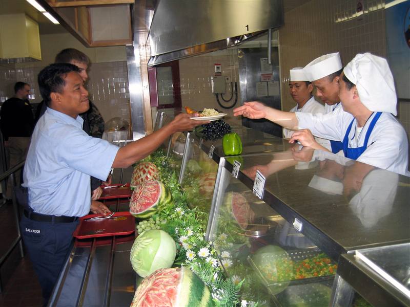 Cooks in white chef uniforms serving a plated meal to service members at a military dining facility